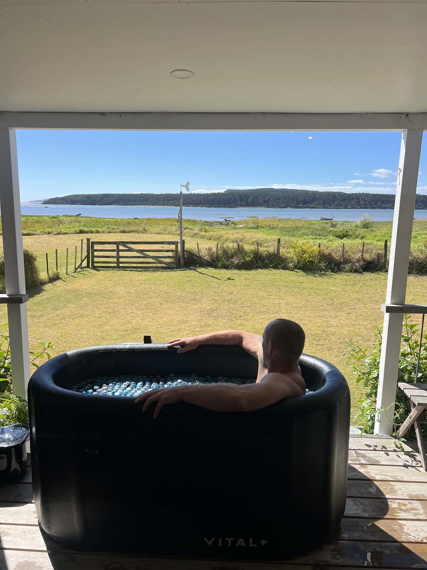 Someone sitting in an ice bath on a deck, looking out over a paddock to the sea